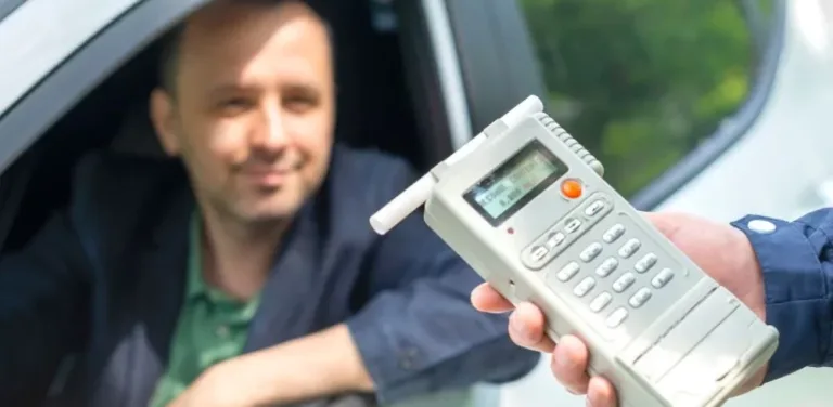 Police officer holding breathalyzer toward driver sitting in car during DUI stop.