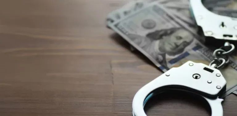Metal handcuffs lying on spread US hundred-dollar bills on a wooden table.