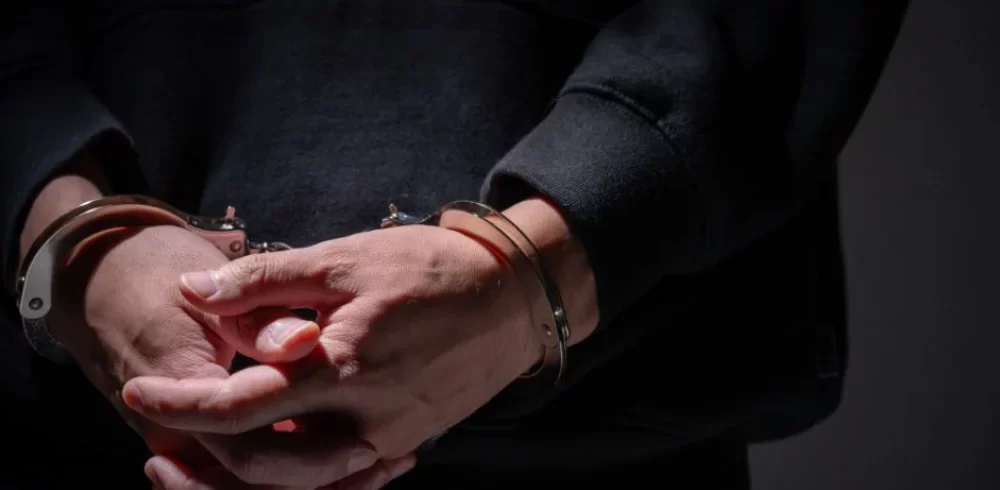 Person in dark hoodie standing with hands cuffed in front in low light.