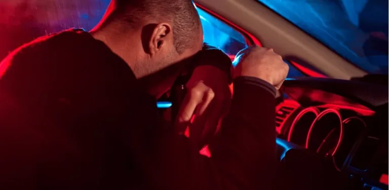 Man slumped over steering wheel in car illuminated by red and blue police lights.