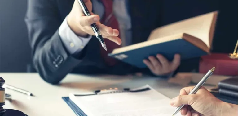 Two professionals in suits review legal documents at a desk with a gavel and an open book nearby.