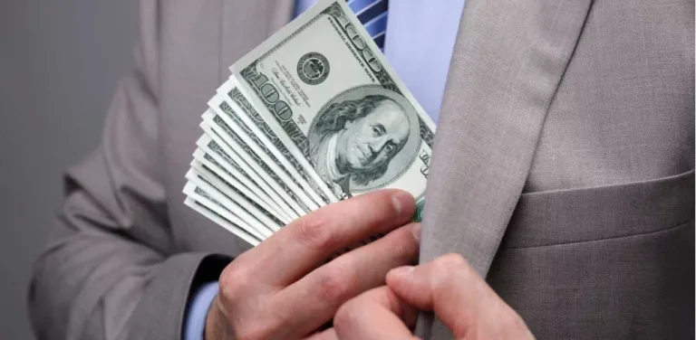 Businessman stuffing a fan of hundred dollar bills into his suit jacket pocket.