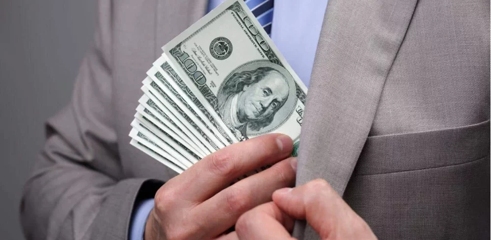 Businessman stuffing a fan of hundred dollar bills into his suit jacket pocket.
