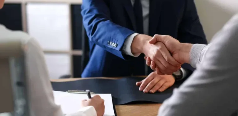 Two individuals in suits shake hands over a wooden desk, signifying an agreement.