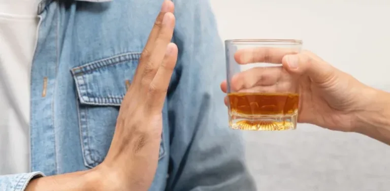 Person refusing offered glass of whiskey with raised hand gesture.