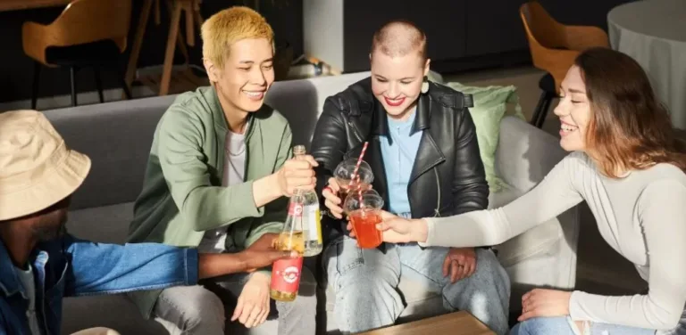 Four friends sitting on a couch clinking bottles and cups at a casual gathering.