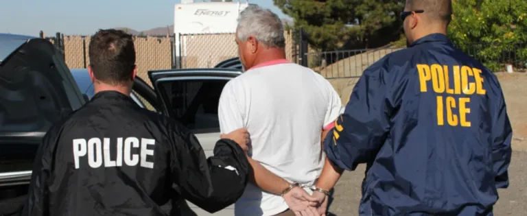 ICE and local police officers escort a handcuffed man toward a vehicle outdoors