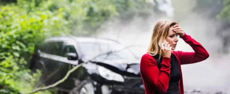 Distressed woman on phone holding her head near a smoking car after an accident in a wooded area