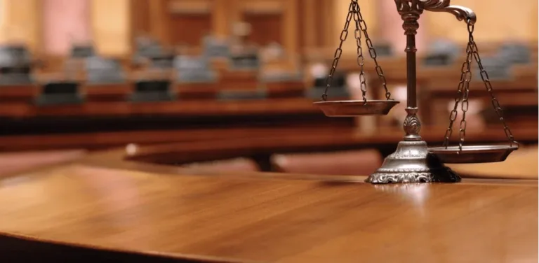 Scales of justice on a wooden courtroom bench with empty seats in the background.