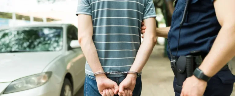 Man in handcuffs standing by a white car with a police officer.